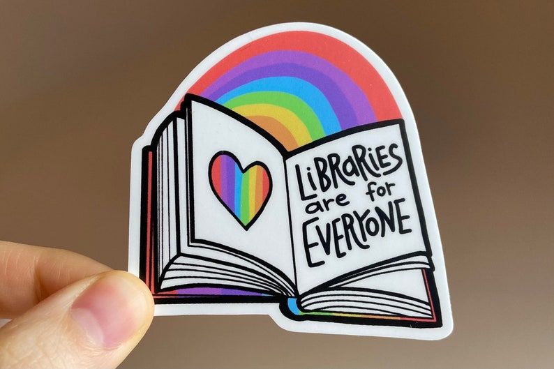 white hand holding a sticker. It's an open book with a rainbow heart on one page and the words "libraries are for everyone" on the opposite. There is a rainbow above the pages. 