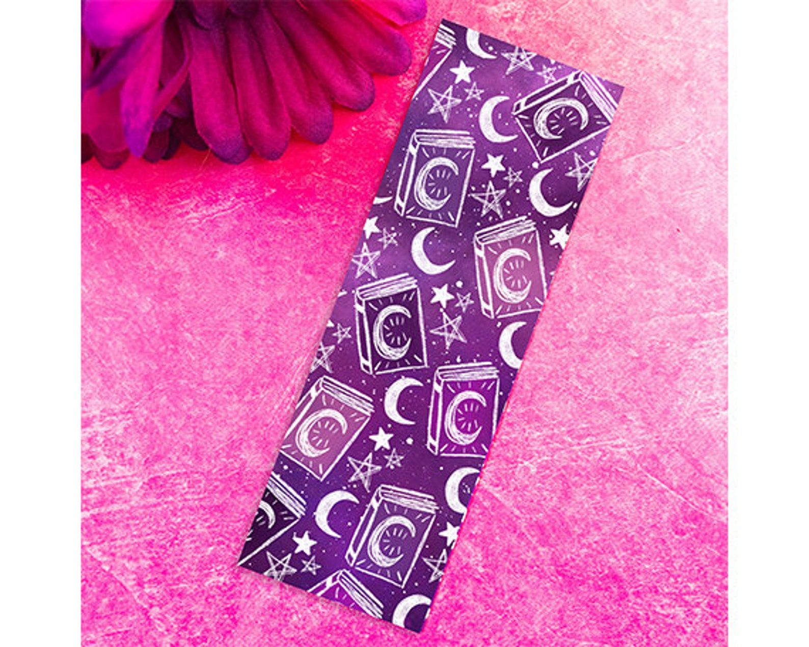 A purple bookmark covered in books, moons, and stars. It sits on a pink background. 
