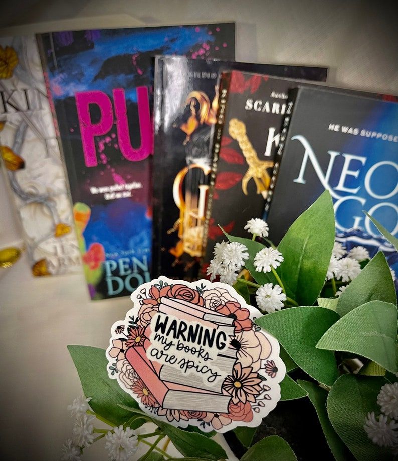 image of a pink sticker on top of a stack of books and some greenery. The sticker reads "warning my books are spicy."