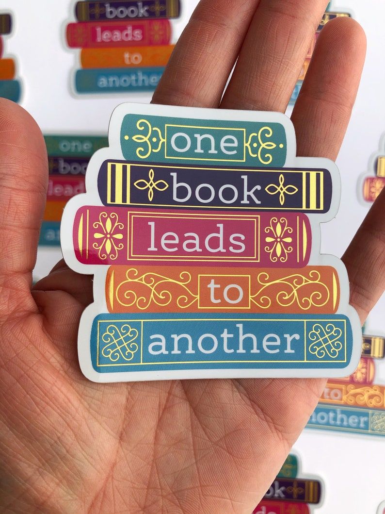 A light brown hand holds a sticker. The sticker is a stack of decorative books with the text "one book leads to another."