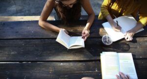 two women are reading or taking notes at a wooden table outside, while a third person reads across the table from them