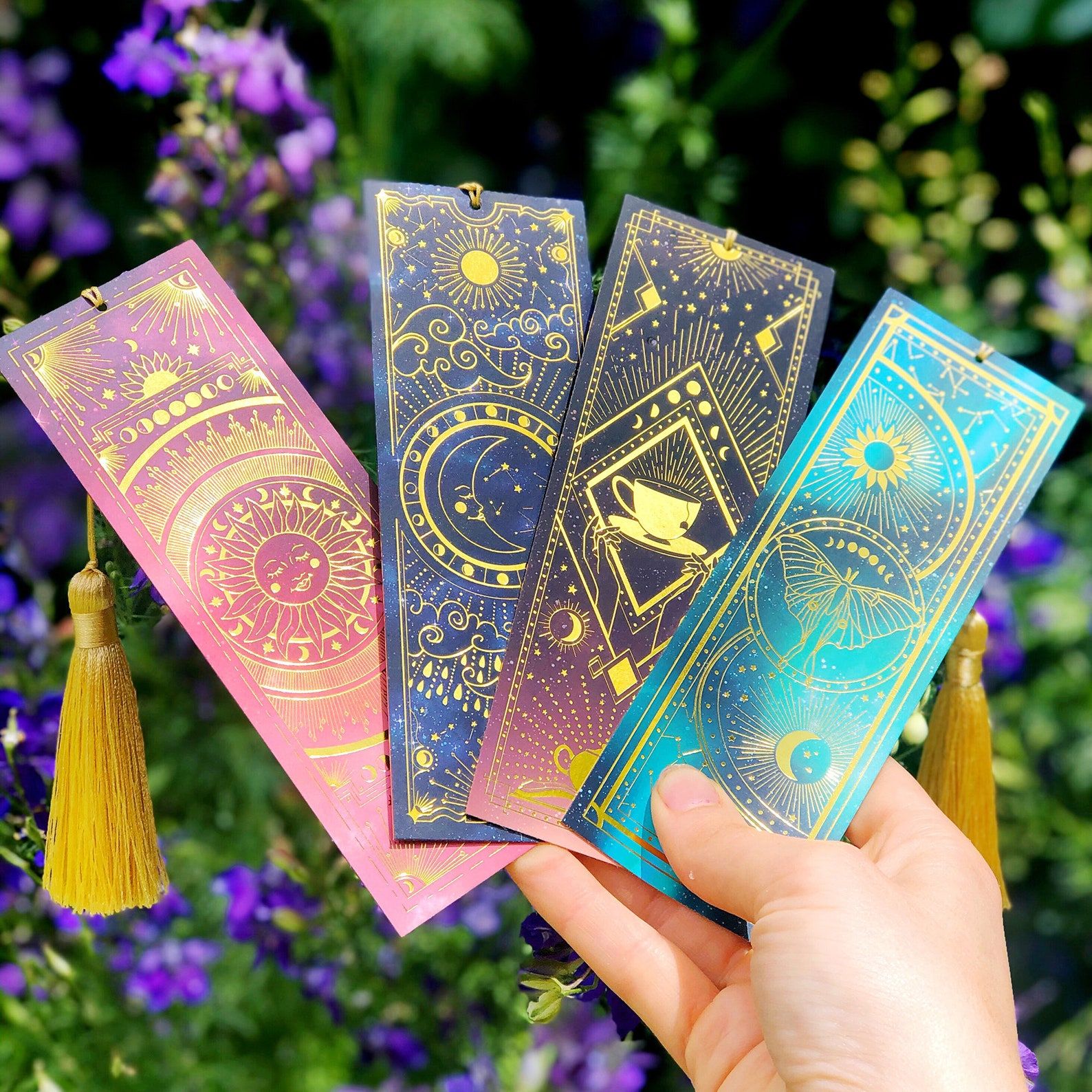 A white hand holds four variations of a brightly colored celestial bookmark. 