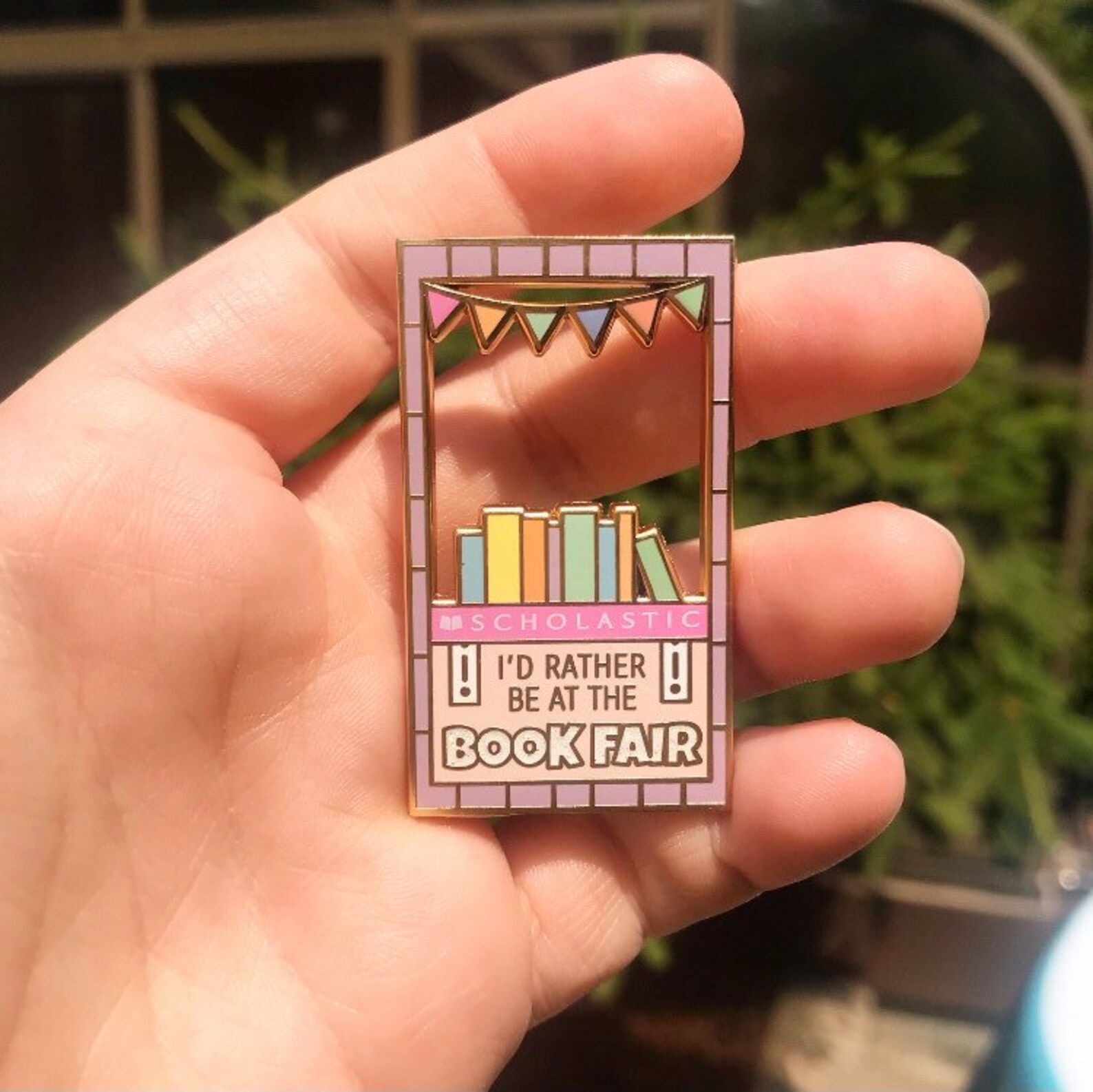 White hand holding a brightly colored enamel pin. It's in the shape of a sales booth and reads "I'd rather be at the book fair," with the Scholastic logo. 