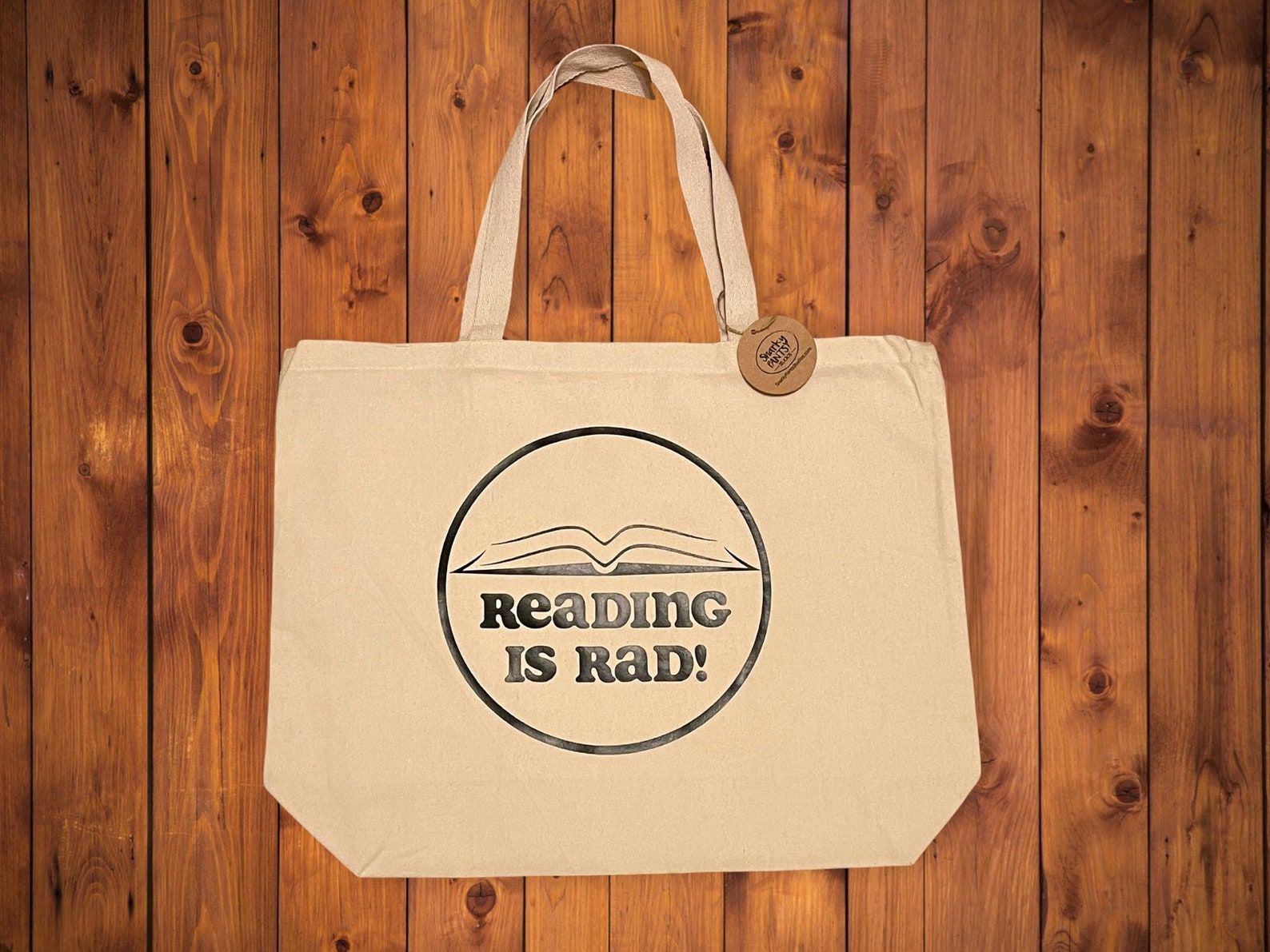 Image of a large canvas tote on a wood background. The center of the tote has a black image of an open book inside a circle. The text beneath the book reads "Reading Is Rad!"