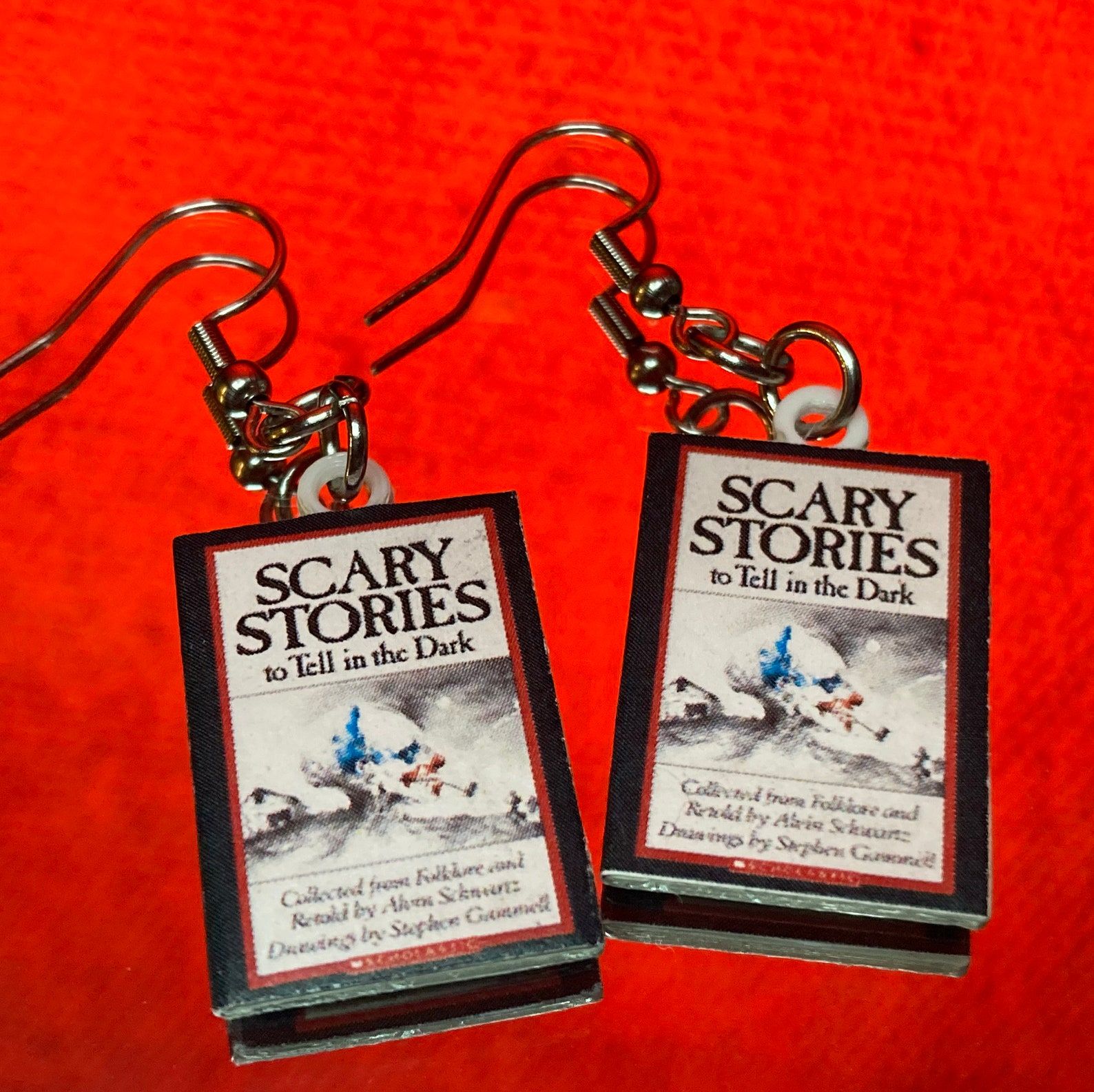 Two dangle earrings on a red background. The earrings are miniature covers of Scary Stories to Tell In The Dark. 