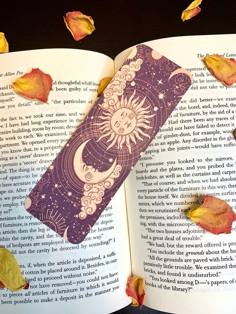 Image of a purple bookmark with cream colored stars, a moon, and a sun inside an open book. There are flower petals on the book, too. 