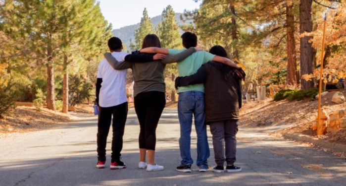 four teens with their backs towards the camera standing on a road in a wooded area with arms linked