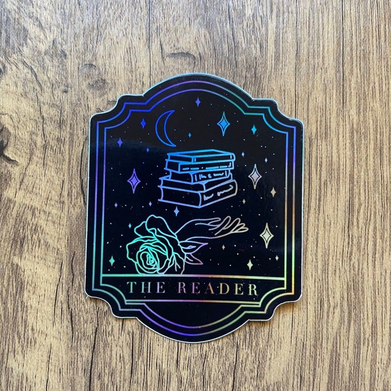 Image of a black sticker on a wood background. The sticker looks like a tarot card called "the reader" and is holographic. 