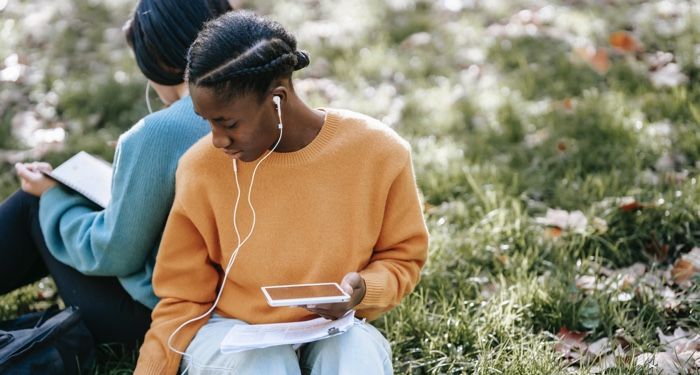 two women wearing headphones, back to back with each other; sitting in the grass. One has a tablet in her lap, and the other a notebook