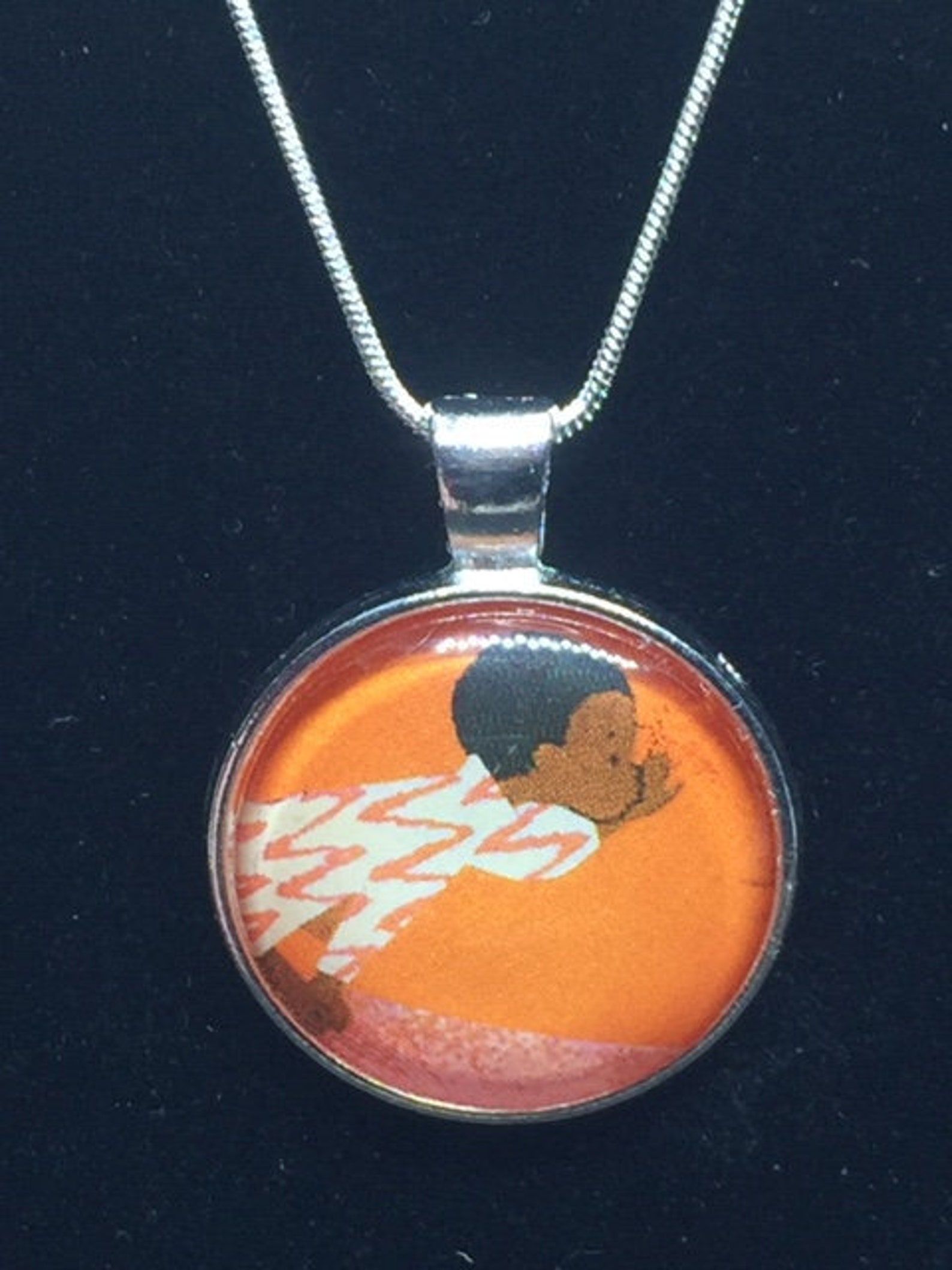 An orange image background with a Black boy from the book A Whistle for Willie made into a necklace pendant. It is on a black background. 