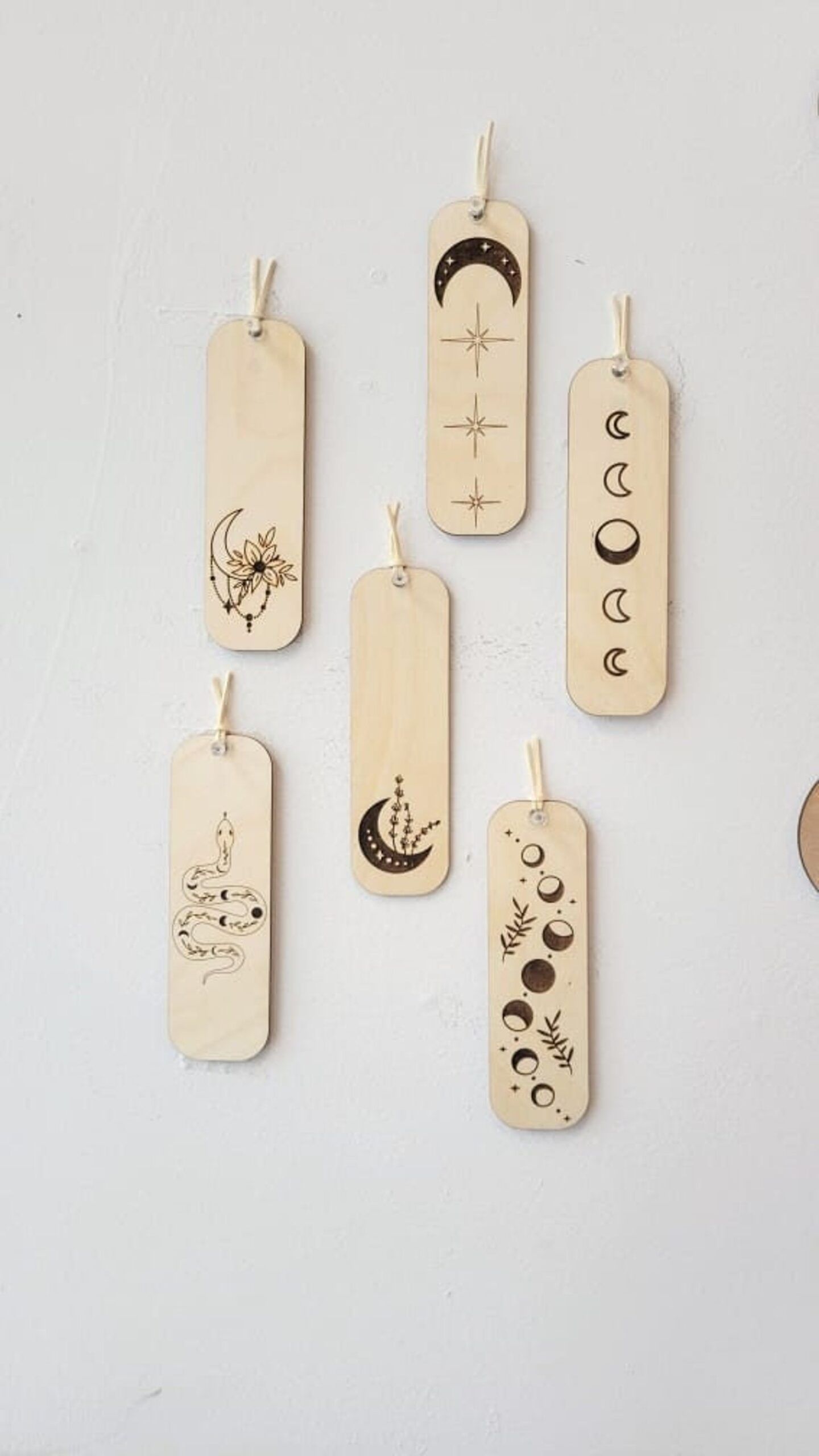 Six staggered wooden bookmarks on a white background. Each bookmark has a celestial theme, including moon phases, moons with flowers, exploding stars, and a starry serpent. 