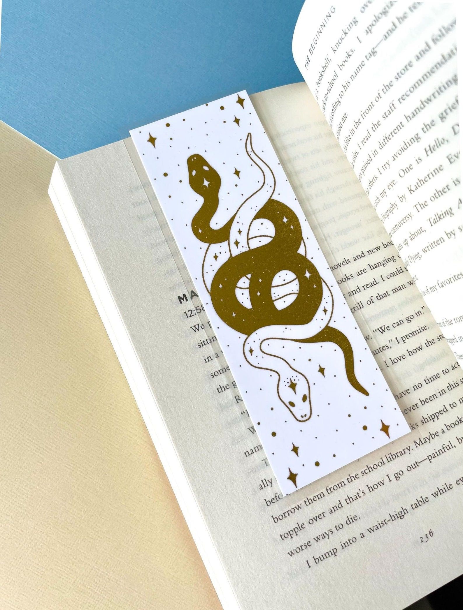 Image of a white bookmark inside a book. The bookmark features two intertwined serpents, one gold and one white, among stars. 