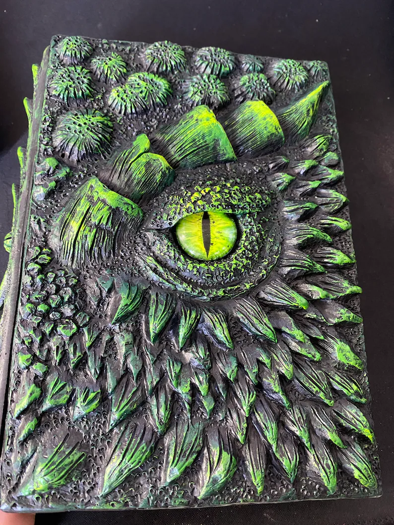 Etsy: A dark gray/black notebook with greenish details on its raised scales and one bright green eye.