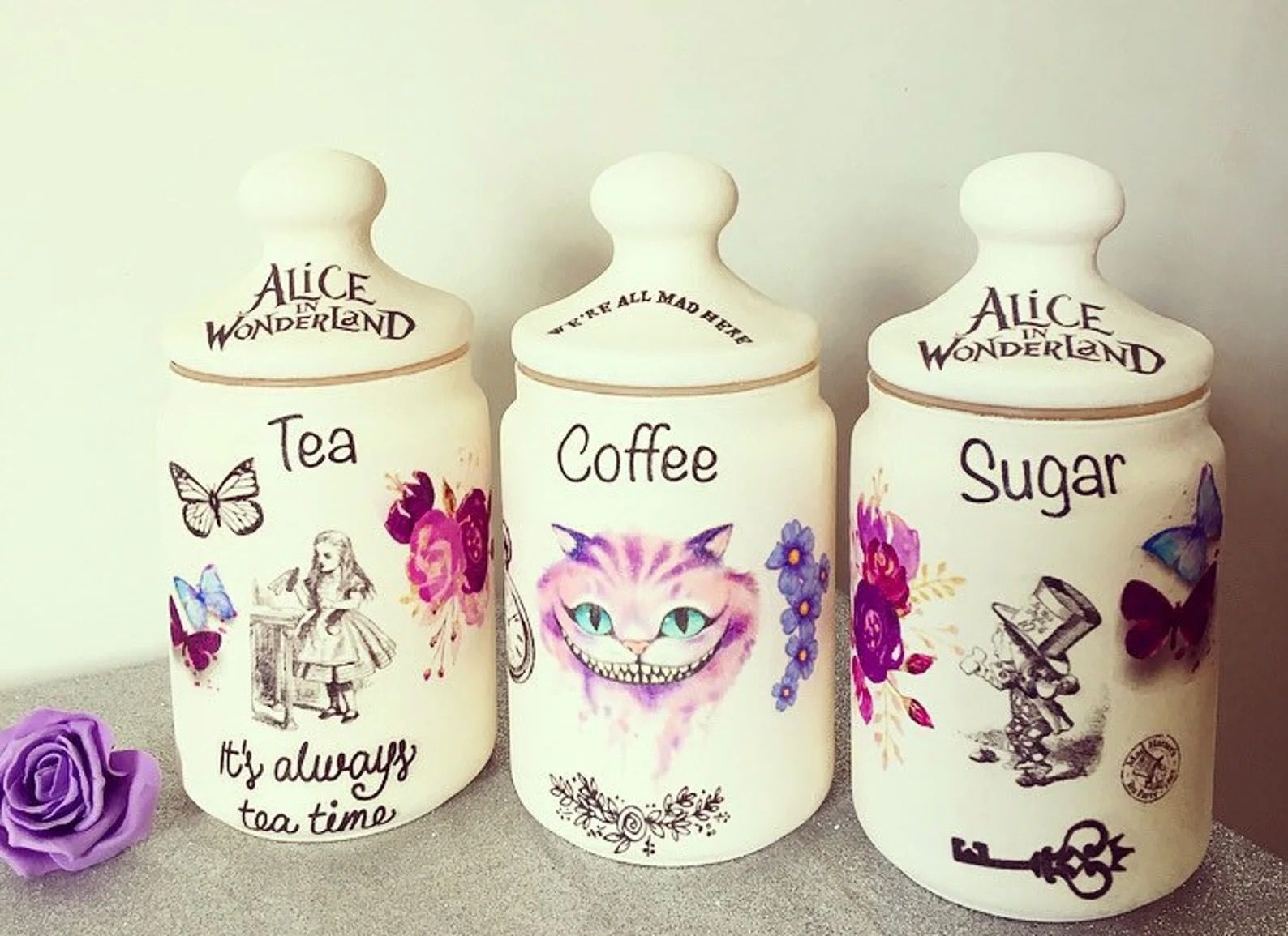 Three white ceramic crocks labeled for tea, coffee, and sugar. Each one has different images from Alice in Wonderland, including the mad hatter, the Chershire cat, and Alice.
