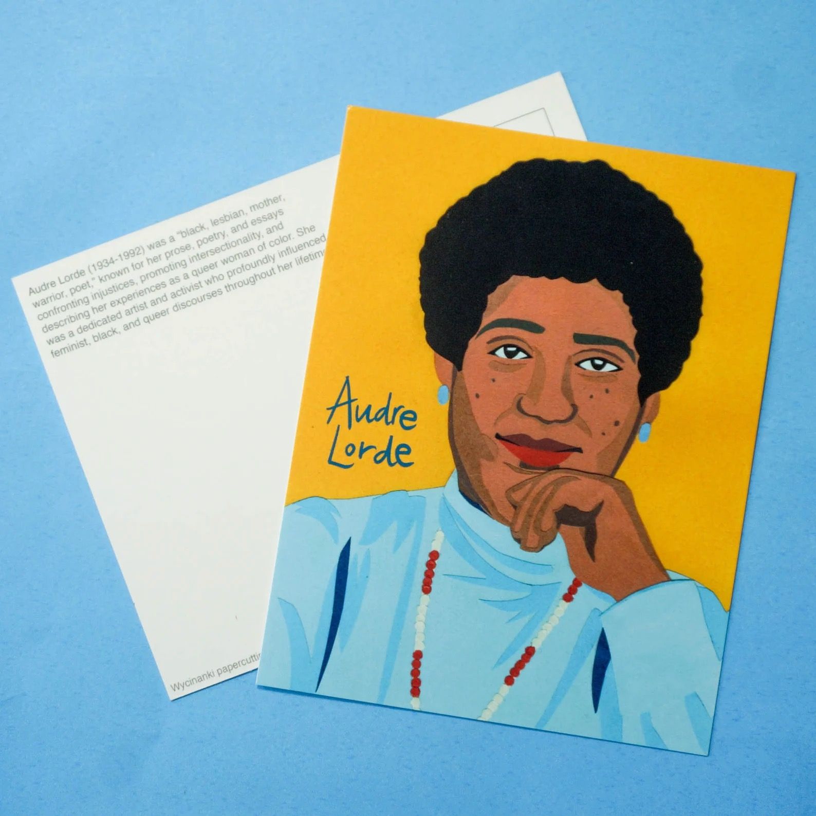 Postcard with a bright painted portrait of Audre Lorde. She has her hand on her chin, and she's wearing a red and white beaded necklace. The background is deep gold.