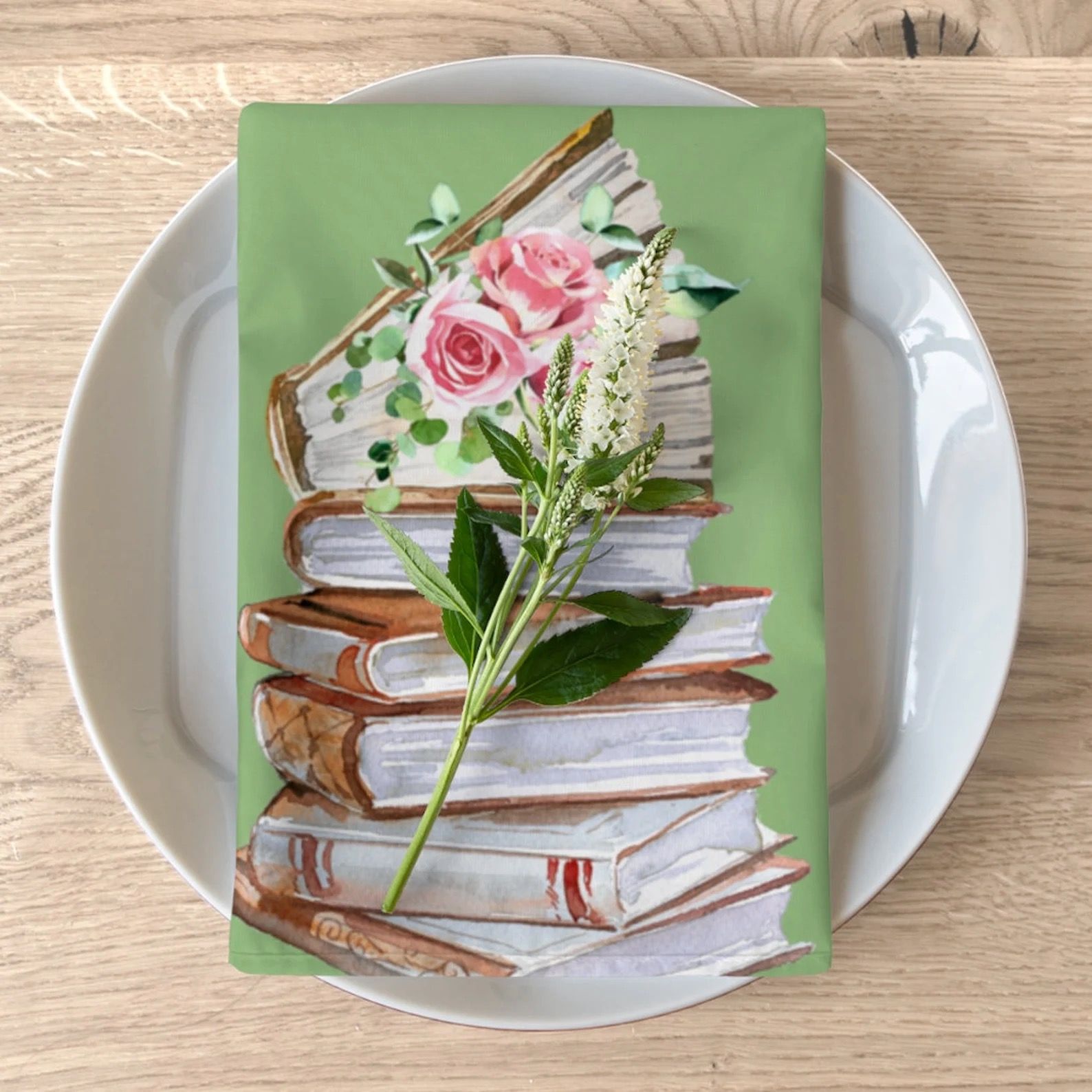 A green cloth napkin is folded on top of a white ceramic plate. The napkin has an image of a stack of old-fashioned leather-bound books, topped with a red rose blossom.