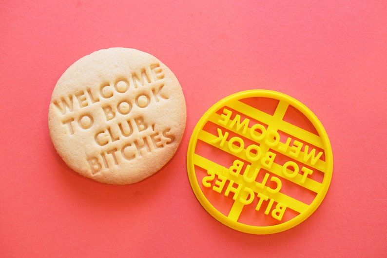 A yellow cookie cutter with a cookie beside it. The cutter and cookie read "welcome to book club, bitches." They're on a pink background. 
