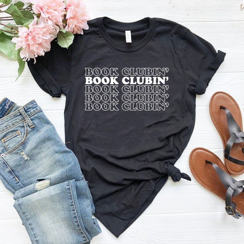 Image of a black t-shirt with the words "book clubbin" down the center, repeating. The shirt is on a white background, with a pair of jeans and sandals beside it. 