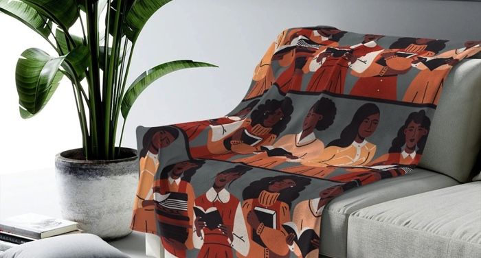 a blanket draped over a grey cough with a large plant off to the left side. The blanket's pattern is a collage of images of Black women reading