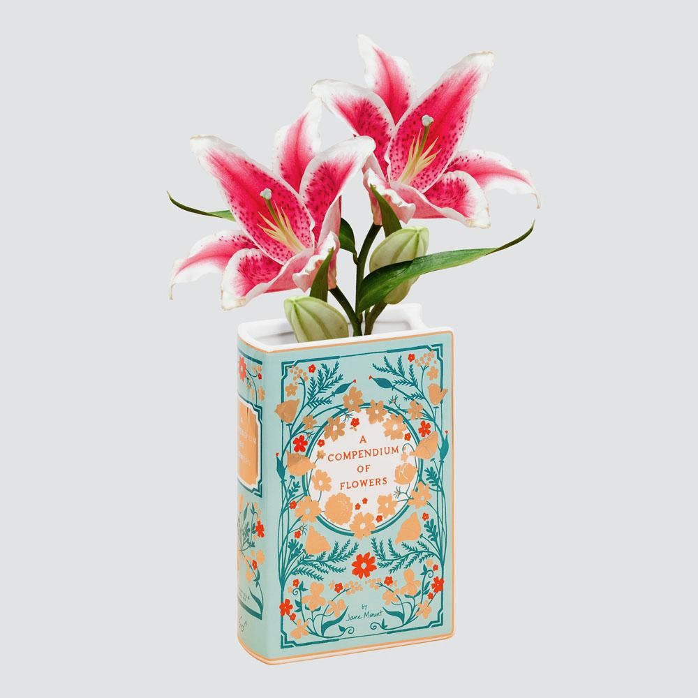 A ceramic vase in the shape of an upright book titled "A Compendium of Flowers" and decorated with a floral vine pattern. The vase holds two lilies.