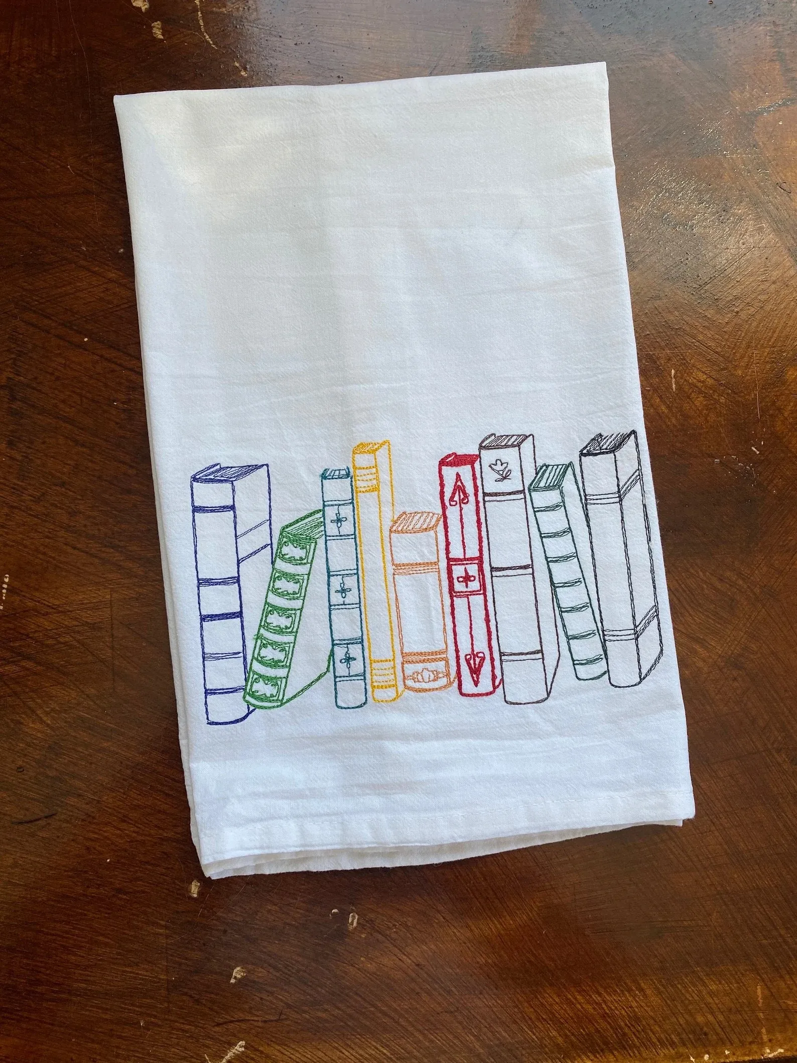 A white dish towel with an embroidered image of a row of books in rainbow colors.