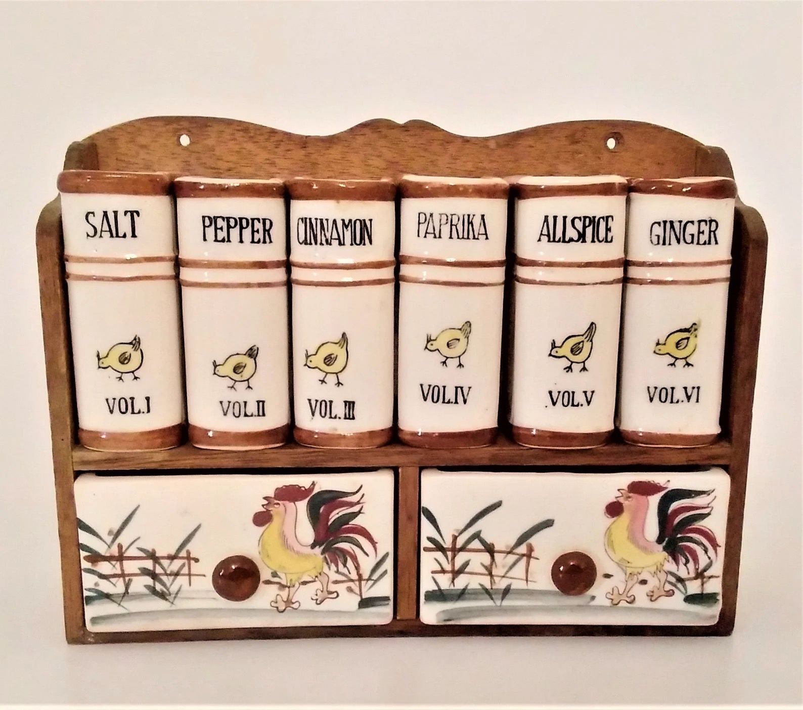 A vintage spice rack holding six ceramic spice jars shaped like books, labeled with the names of spices and 'Vol I, Vol II, Vol III' etc. The shelf has two small drawers painted with roosters below the spice jars.
