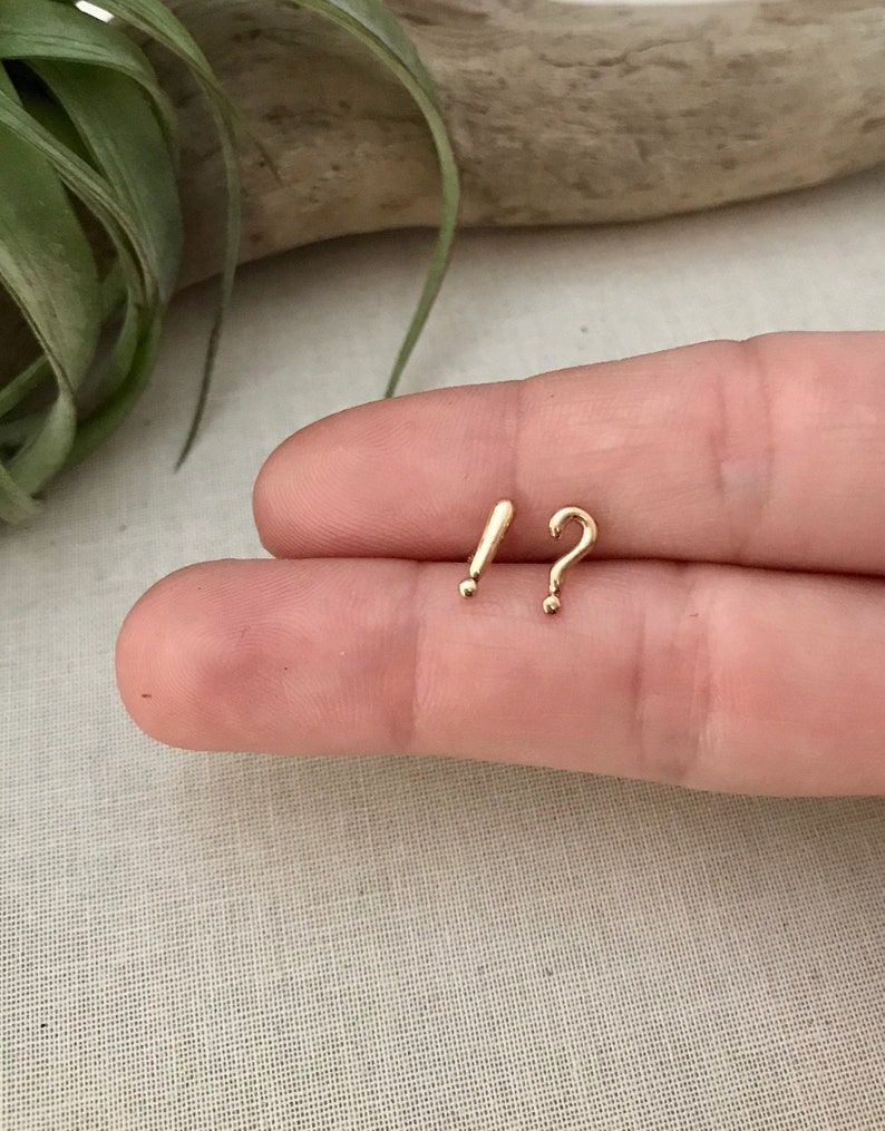 Small gold earrings in shape of question mark and exclamation mark. They're on a white hand. 