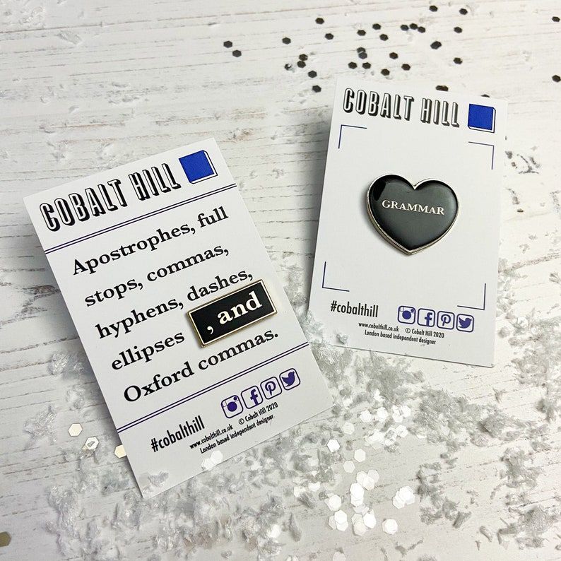 Set of two enamel pins. One is of the Oxford comma and one is a heart with the word grammar inside. Both are black with white font. 