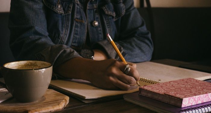 a brown-skinned person wearing a denim jacket writes in a journal with a pencil. There is a cup of coffee and another book on the table.