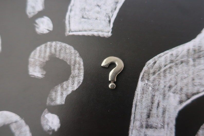 A pewter question mark pin on a background made of black with white chalk question marks.
