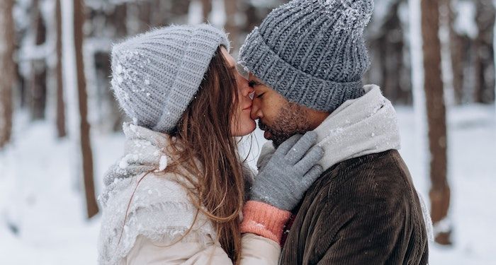a photo of a couple outside by a forest in the snow. A white woman kisses a Black man on the nose.