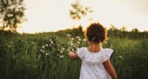 the back of a small child with brown skin picking flowers in a meadow