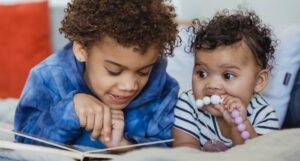 a photo of two Black children reading a board book together. One is a toddler chewing on a teething necklace