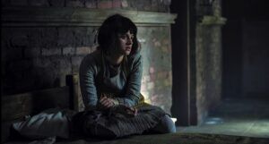 Anya Chalotra as disabled character Yennefer in a still frame from Netflix series The Witcher. Character is sitting with crossed arms before a brick hearth