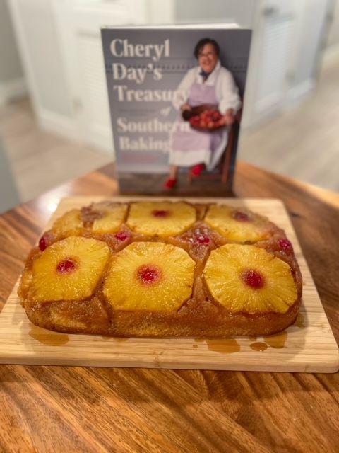 Image of a rectangular pineapple upside-down cake with six pineapple rings and maraschino cherries on top, sitting on a wooden cutting board in front of Cheryl Day's Treasury of Southern Baking cookbook