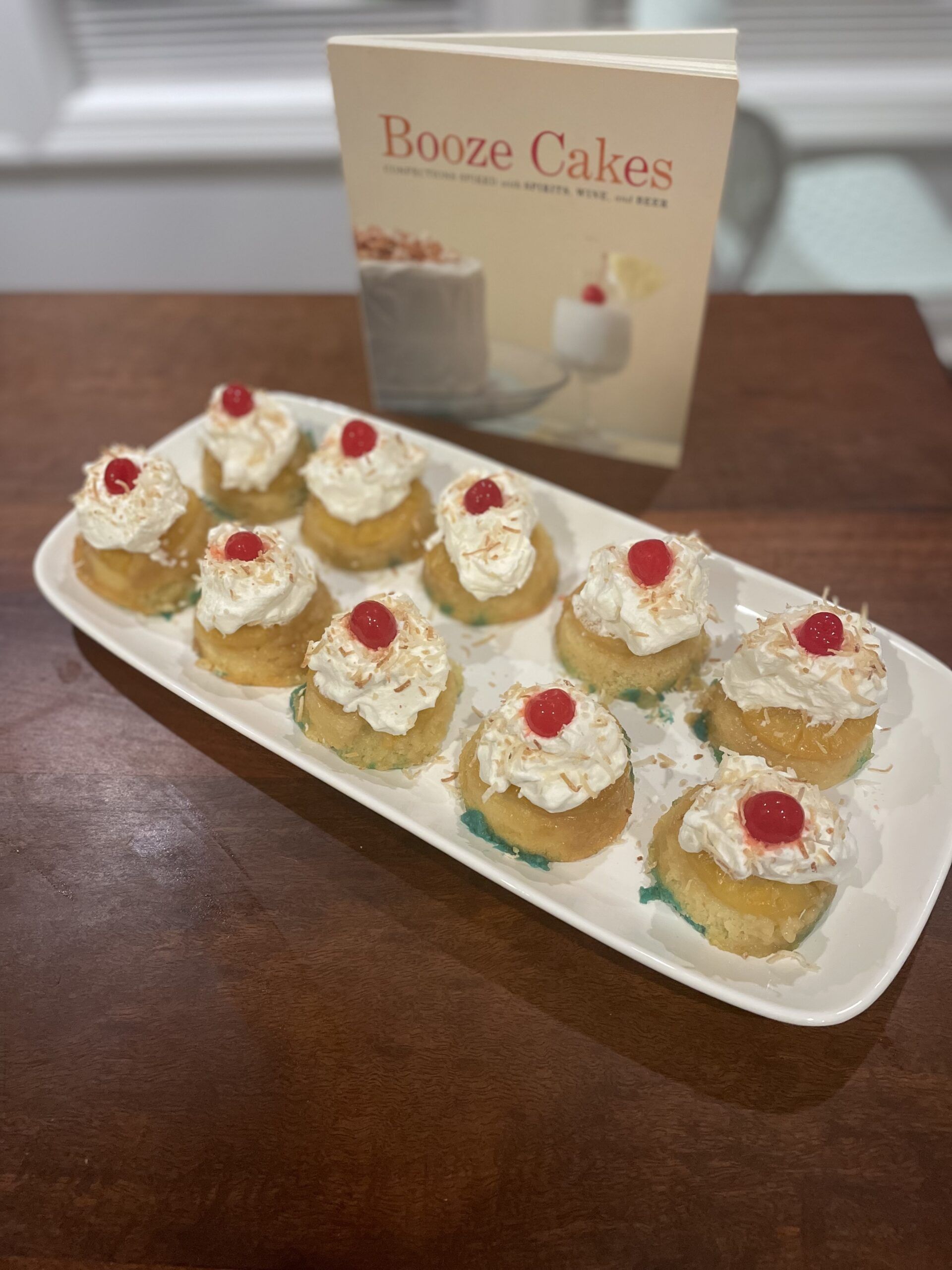 Image of twelve cupcakes on a rectangular serving dish. Each cupcake is topped with a pineapple ring, whipped cream, cherry, and toasted coconut. The tray sits in front of Booze Cakes cookbook.