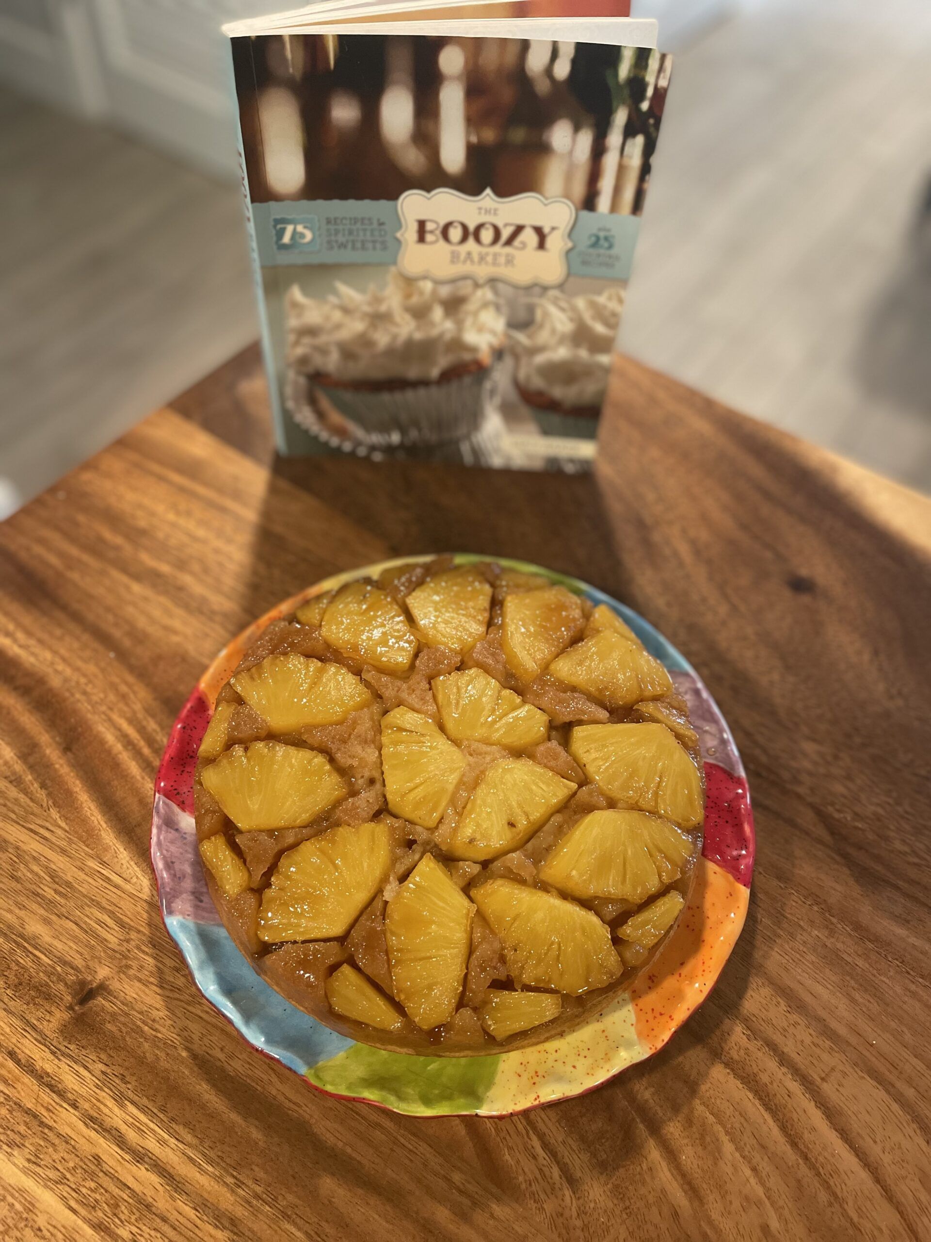 Image of a round pineapple upside-down cake with chunks of fresh pineapple on top. The cake is on a rainbow cake plate in front of The Boozy Baker cookbook.