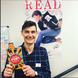 a photo of author Mitch Johnson holding up the book Pop! in front of a library READ sign