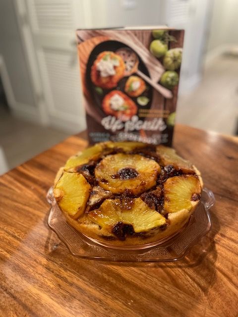 Image of a round pineapple upside-down cake with a dark walnut sauce on a glass cake plate in front of The Up South Cookbook