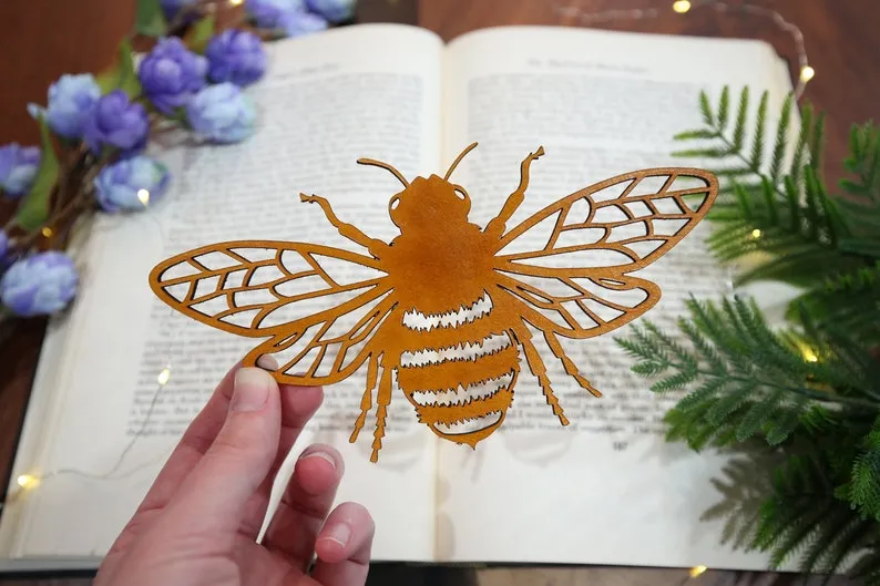 Image of a white hand holding a leather, laser-cut bookmark in the shape of a bee. 