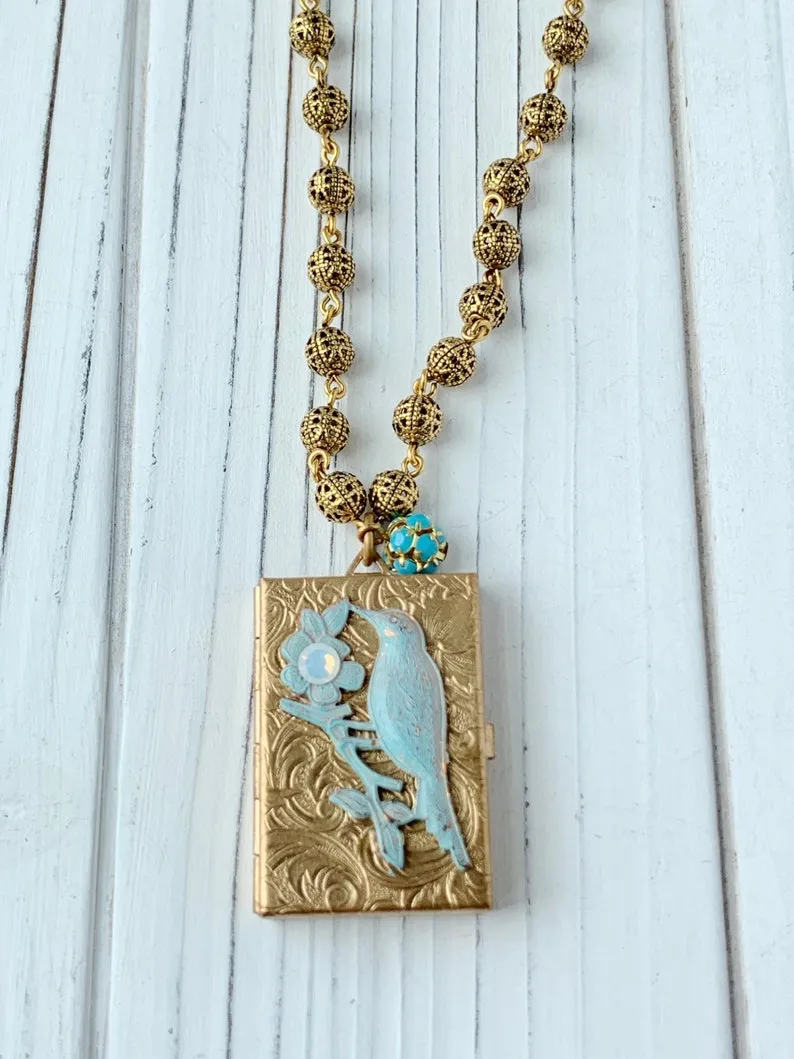 Image of a gold plated locket with a blue bird on it. The locket is designed to look like a vintage book. 