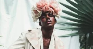 man with dark brown skin wearing a pink rose crown and white clothing posing for the camera