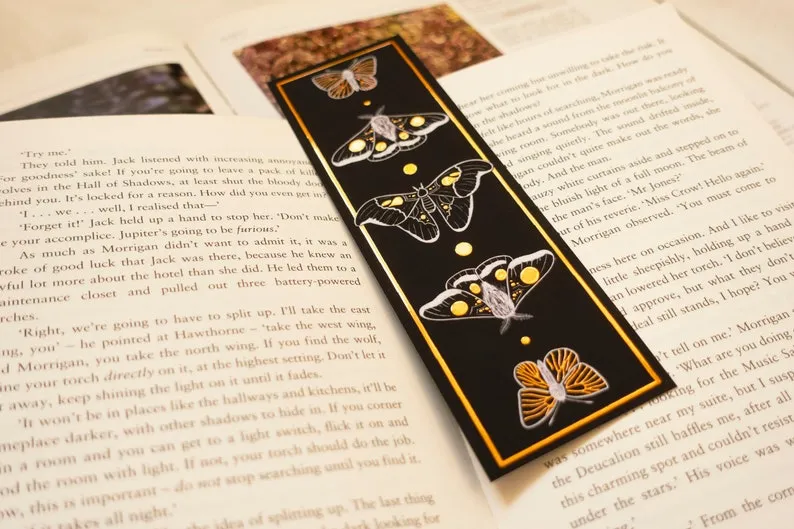 Image of a black bookmark featuring gold foil moths in the center of an open book.