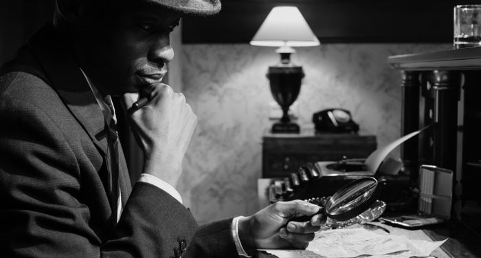 black and white photo of a Black detective looking through a magnifying glass