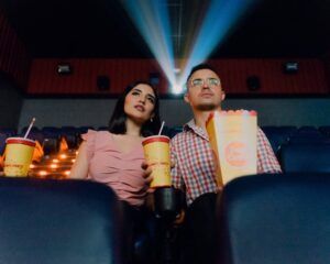 a light-skinned man and woman sitting next to each other with drinks and popcorn in a dark movie theater
