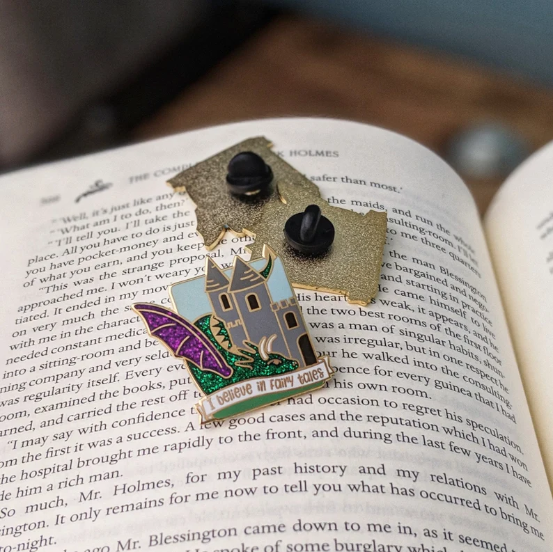 Image of the front and back of an enamel pin on an open book. The pin reads "I believe in fairy tales" and includes a dragon and a castle. 