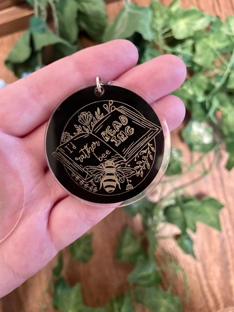 Image of a white hand holding an acrylic keychain. It has a black background and gold coloring. The keychain features an open book and a bee and reads "I'd rather bee reading."