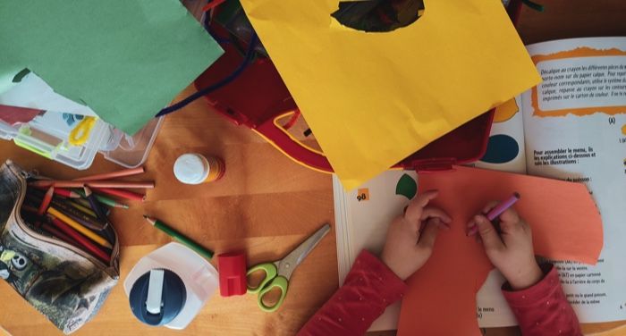 hands of a child using a colored pencil to draw Ono construction paper with an open book beside them, as seen from above