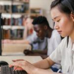 a person working at a desktop computer in a library, two more people doing the same in the background