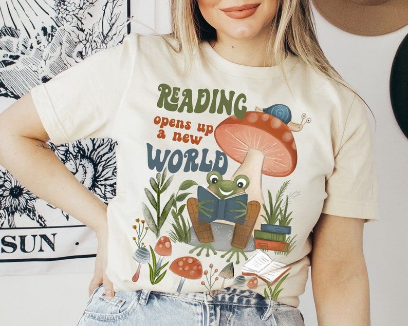 Image of a white person wearing a cream colored shirt. The shirt has a frog reading beneath a toadstool. It's a retro color scheme and has in retro font the words "reading opens up a new world."